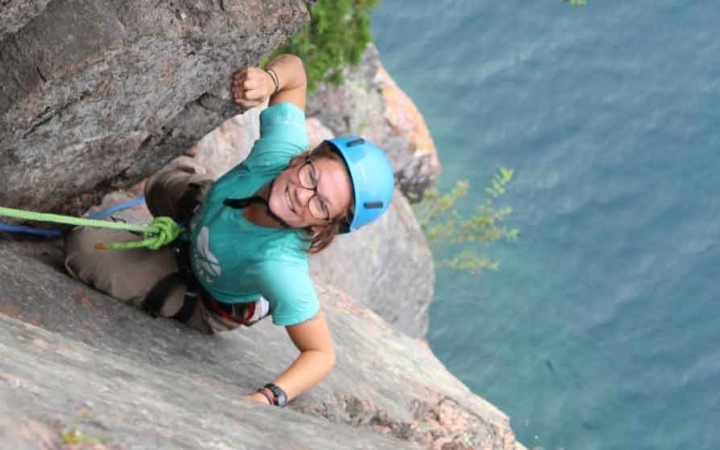 a student who is rock climbing above a body of water smiles at the camera